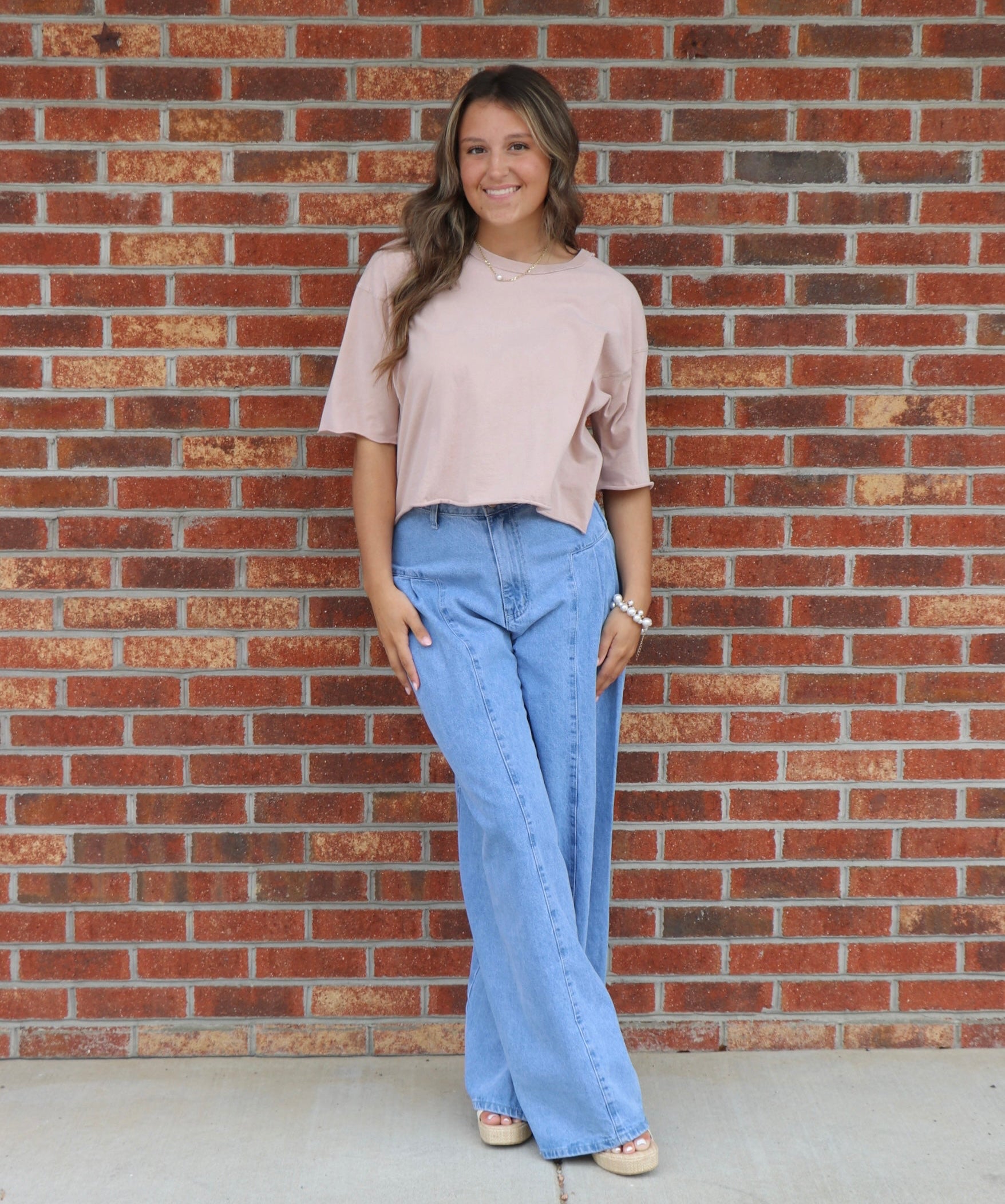 Woman wearing a beige top and blue jeans standing in front of a brick wall.