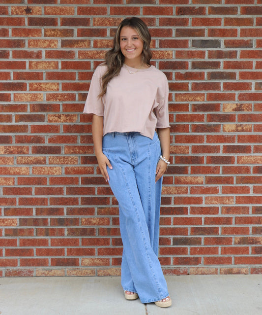 Woman wearing a beige top and blue jeans standing in front of a brick wall.