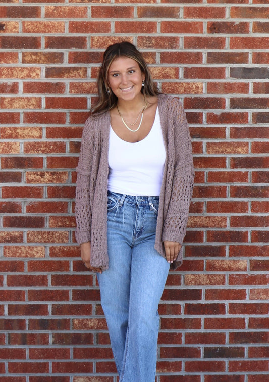 Woman wearing a cardigan, white top, and blue jeans standing against a brick wall.